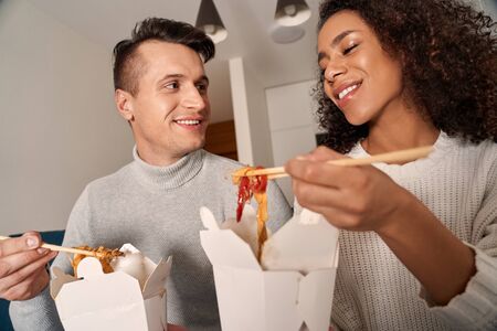 Romantic Date. Young multiethnic couple at home sitting on sofa eating noodles talking smiling joyful close-upの写真素材