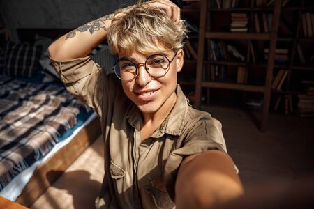 Blogger. Young woman short hair in glasses sitting at home recording vlog on phone smiling happy close-upの写真素材