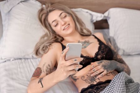 Weekend at Home. Young tattooed woman in lingerie lying on bed at bedroom browsing internet on smartphone close-up smiling relaxedの写真素材