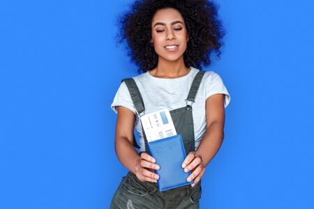 Tourism. Mulatto girl wearing denim overall standing isolated on blue with passport and ticket close-up smiling happy blurred backgroundの写真素材
