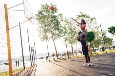 Outdoors Activity. Girlfriend sitting on shoulders of boyfriend standing on basketball court throwin...
