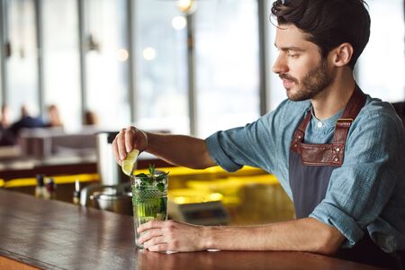 Making Drink. Bartender standing at counter decorating glass of mojito with lime smiling satisfiedの写真素材