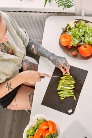 Leisure at Home. Young tattooed woman standing at kitchen cutting vegetables cooking meal top view close-upの写真素材
