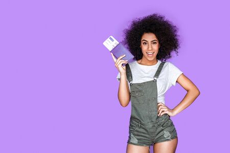 Tourism. Mulatto girl wearing denim overall standing isolated on blue with passport and ticket posing to camera cheerfulの写真素材