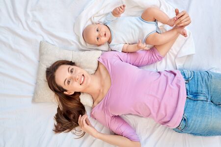 Young adult mom looking at camera, lying on bed with little newborn offspringの写真素材