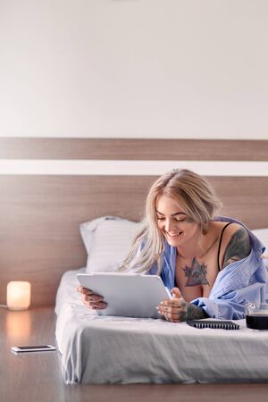 Weekend at Home. Young tattooed woman in lingerie lying on bed at bedroom watching video on digital tablet smiling cheerful drinking coffeeの写真素材