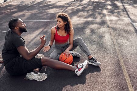 Outdoors Activity. African couple sitting with ball on basketball court discussing game top view