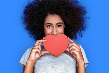 Valentines Day. Mulatto girl standing isolated on blue covering face with valentine playful close-upの写真素材