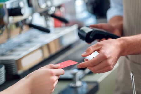 Cropped view of young adult barista working in cafe, holding wireless terminal and taking card from client to making payment for coffeeの写真素材