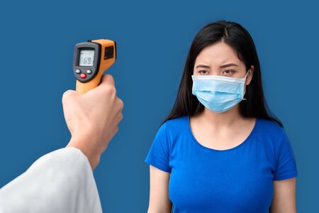 Coronavirus Concept. Doctor with thermal camera against unhappy chinese woman in medical face mask...
