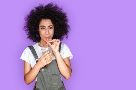 Freestyle. Mulatto girl wearing denim overall standing isolated on blue drinking smiling playfulの写真素材
