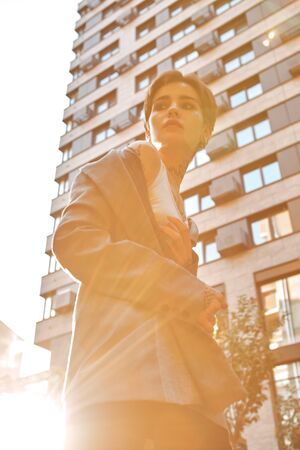 Urban Style. Young stylish woman in coat standing outdoors looking aside pensive in sunlight bottom viewの写真素材