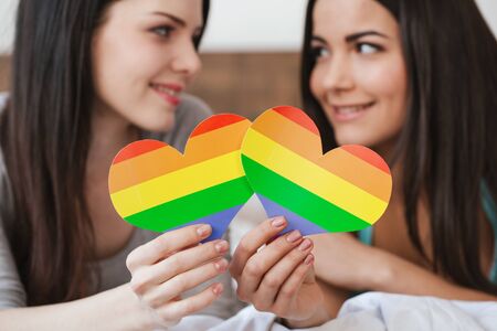 Lesbian couple in bedroom at home lying holding card lgbt color close-up looking at each other passionateの写真素材