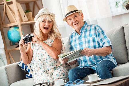 Senior couple sitting in travel agency sitting wearing hats holding camera and map surprisedの写真素材