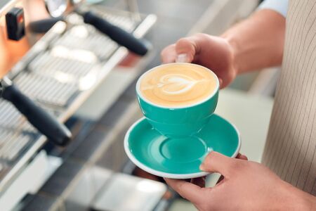 Cropped view of young adult barista working in cafe, standing near professional coffee machine, holding cup of beverage in handの写真素材