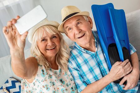 Senior couple sitting in travel agency sitting wearing hats husband holding flippers while wife taking selfie photos on smartphone smiling happyの写真素材