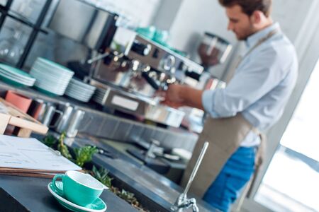 Selective focus on mug with beverage against blurred background with young adult barista working...