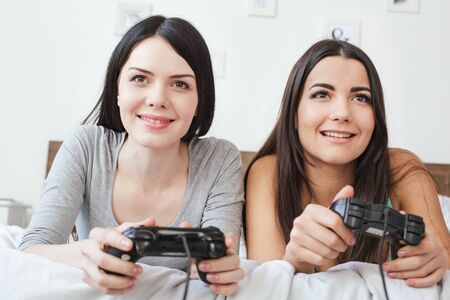 Lesbian couple in bedroom at home lying holding controllers playing game cheerful close-upの写真素材