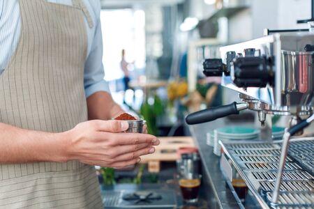 Cropped view of young adult barista in apron working in cafe, standing near professional espresso machine with freshly ground coffee in filterの写真素材