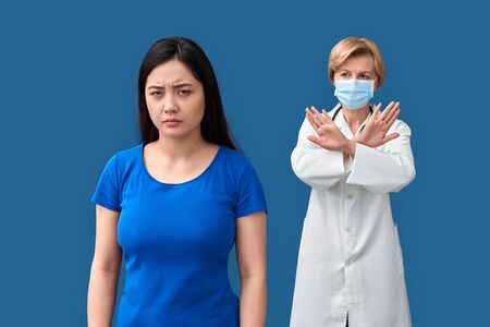 Coronavirus Concept. Doctor in medical mask and chinese woman standing isolated on blueの写真素材
