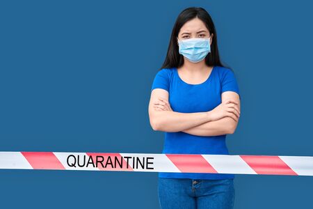 Coronavirus Quarantine. Chinese woman in medical mask standing isolated on blue behind warning tape crossed hands frowning unhappyの写真素材