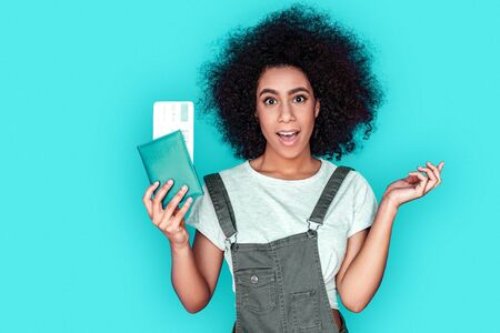 Unexpected trip. African american girl wearing denim overall standing isolated on blue with passport and ticket surprisedの写真素材