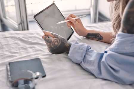 Cropped view of one young adult girl wearing in casual shirt, lying on bed, using modern tablet computer in bedroomの写真素材