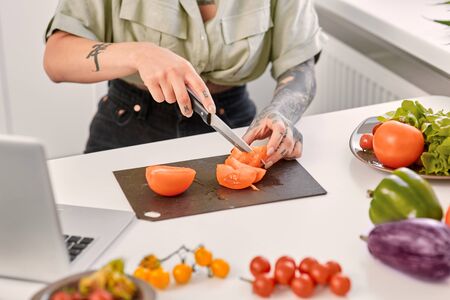 Cropped view of one young adult girl wearing in casual shirt holding knife in hands, cooking on kitchen, cutting tomatoの写真素材