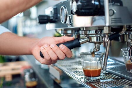 Young handsome man barista standing at coffee shop holding portafilter using machine pouring espresso shot into glass close-upの写真素材