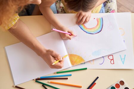 Young mother and daughter education at home sitting at table drawing together using color pencils rainbow and sun positive vibes top viewの写真素材