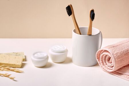 Concept of choosing natural body care products. Two eco and bamboo toothbrush in cup, container with cream, soap and towel on white beige backgroundの写真素材