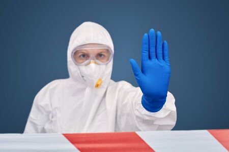 Woman doctor wearing white hazmat suit goggles medical gloves and respirator standing behind red warning tape isolated on blue backround quarantine showing stop gesture close-upの写真素材