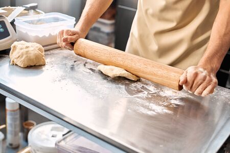 Cropped view of young adult man working with rolling pin and dough on kitchen in bakery manufacturing, making sweet mini croissants and fresh breadの写真素材
