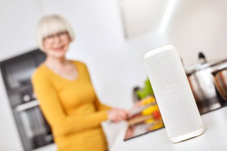 Selective focus of wireless digital assistant telling recipe of healthy food to happy senior woman while she standing in kitchen at homeの写真素材