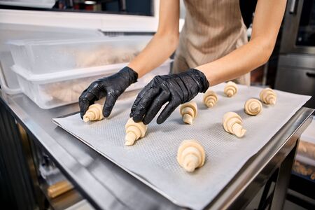 Young woman wearing apron and gloves at bakery shop small business producing croissants arranging pastry on tray close-upの写真素材