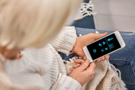 Cropped view of elderly woman using modern smartphone with smart house app, resting at home, sitting...