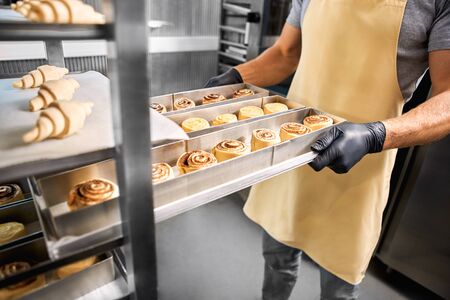 Young man wearing gloves standing at bakery shop small business holding tray of cinnamon rolls...