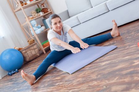 Healthy Lifestyle. Young woman doing yoga at home sitting legs apart bending down stretching smiling...