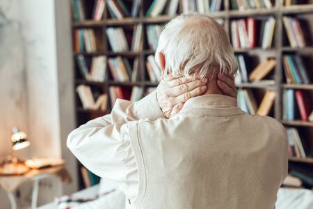Senior man alone standing at library massaging neck back view close-upの写真素材