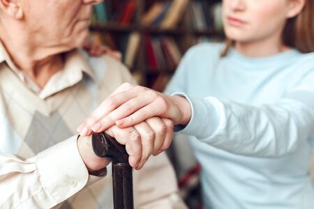 Senior man and young woman at home library sitting on sofa father leaning on walking stick daughter holding his hand supportive close-upの写真素材