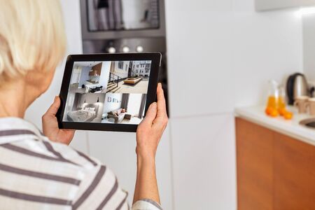 Senior woman at home standing at kitchen holding digital tablet controlling smart home system back view checking cameras close-upの写真素材