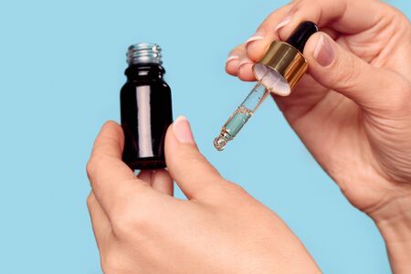 Skincare concept. Cropped view of middle aged female holding glass bottle with organic anti aging oil, applying serum on skin, standing isolated on pastel blue background with copy spaceの写真素材