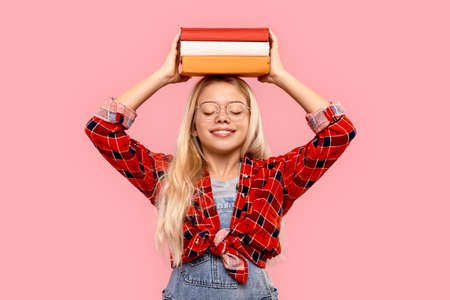 Freestyle. Little girl in eyeglasses standing isolated on pink with books on head closed eyes smiling joyfulの写真素材
