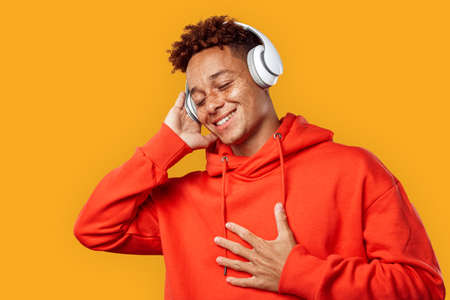 Freestyle. Mulatto freckled guy in hoodie and headphones standing isolated on yellow listening music smiling delightfulの写真素材