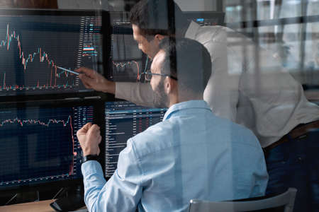 Stock Traiding. Traders sitting at office monitoring price flow analyzing chart talking excited through glass shotの写真素材