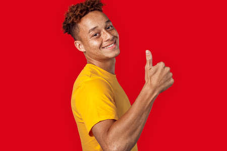 Freestyle. Mulatto freckled guy in yellow t-shirt standing isolated on dark pink showing thumb up smiling confidentの写真素材