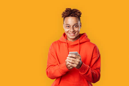 Freestyle. Mulatto freckled guy in hoodie standing isolated on yellow browsing internet on smartphone happyの写真素材