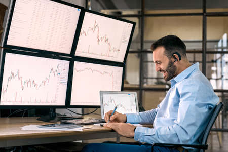 Stock Traiding. Trader wearing headset sitting at office in front of monitors with data chart doing online trading training writing down key pointsの写真素材