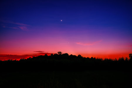 Shadows of forests and hills under the moon and stars contrasted with color red and blue of night sky.の写真素材