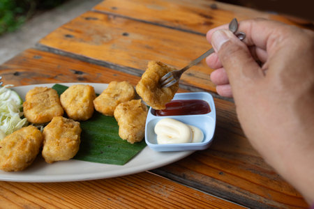 Someone hand uses fork to get a picec of chicken nuggets from disk on wooden table.の写真素材